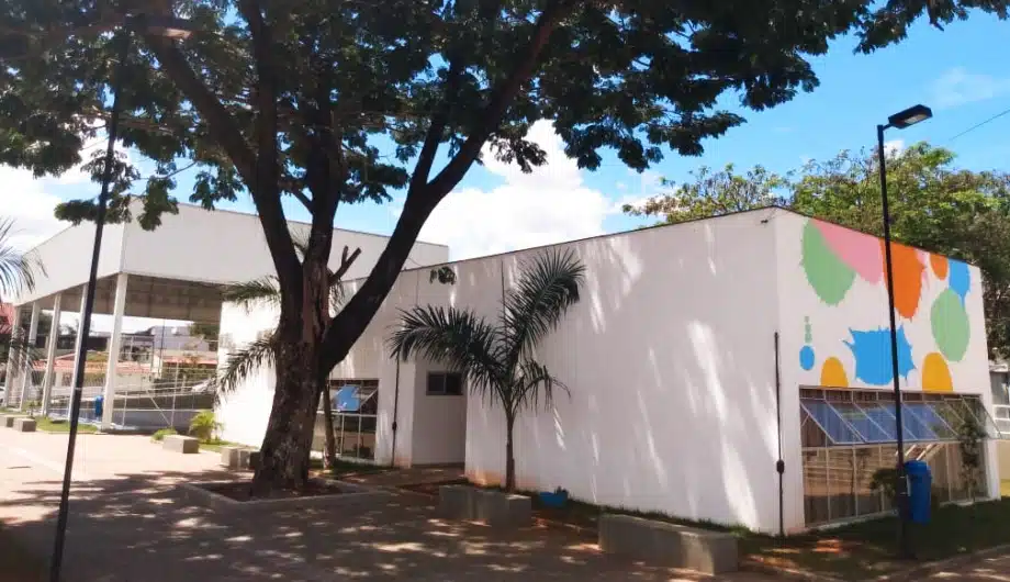 Temos a visão do prédio da biblioteca Sergio Sampaio, bem atrás de uma arvore. O prédio é da cor branca com detalhes coloridos na lateral do prédio.