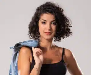 Gabriela posa com uma blusa preta de alça fina, e segurando uma jaqueta sob os ombros. A humorista olha para a câmera.
