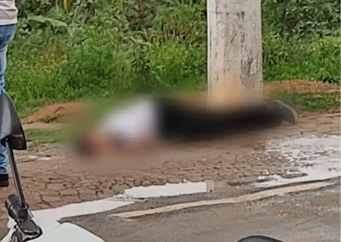 Imagem retrata homem caído vítima de disparo de arma de fogo. A imagem está embaçada. A vítima veste blusa branca e calça preta.