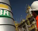 Petrobras anuncia redução de 5,6% no preço da gasolina Homem usano uma blusa vermelha e capacete branco, olha para um tanque com a logo da Petrobras