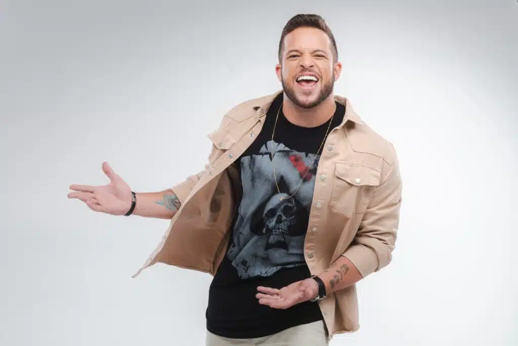 Flavio Andrade posa usando uma camisa preta de estampa e uma calça creme. Por cima, usa uma jaqueta de outra textura.