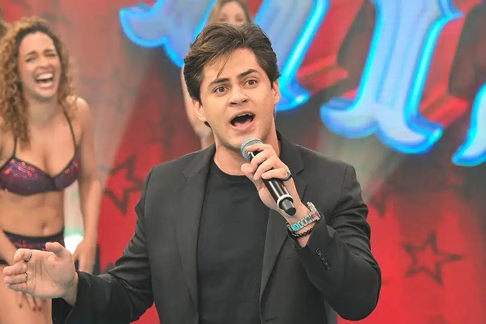 O comediante na imagem aparece falando no microfone, Lucas usa uma camisa e blase preto.