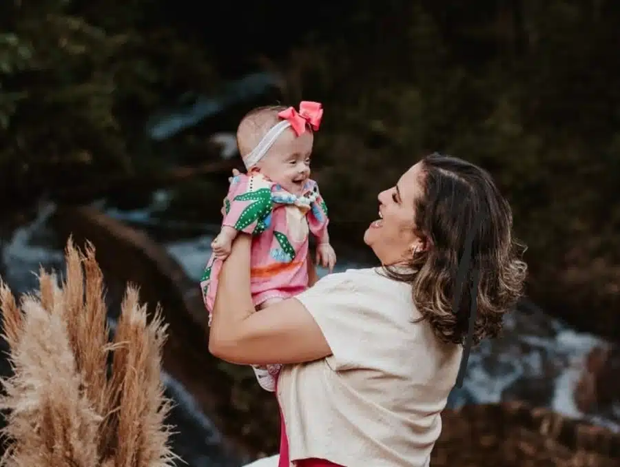 Uma mãe segura bebê no alto com as mãos,olha para ela sorrindo. A criança também olha para a mãe e sorri.