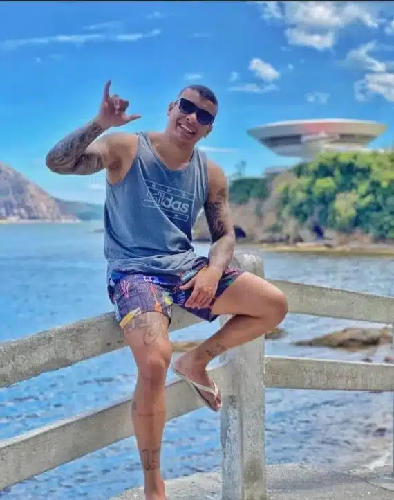 rapaz de short e camisa regata, sentado em uma cerca próximo ao mar