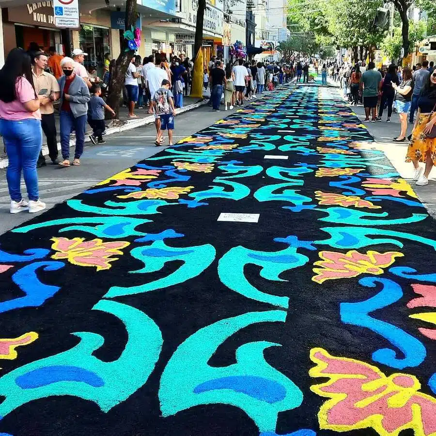 Imagem de um dos tapetes da festa, posta em uma passarela. As pessoas passam e olham para a tapeçaria.