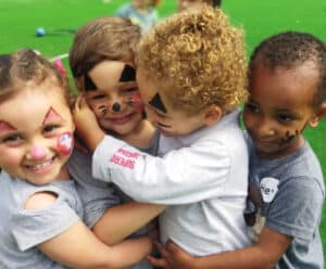 Quatro crianças se abraçamdo em um campo verde. Seus rostos estão pintados como de uma palhaço e sorridentes.