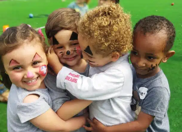 Quatro crianças se abraçamdo em um campo verde. Seus rostos estão pintados como de uma palhaço e sorridentes.