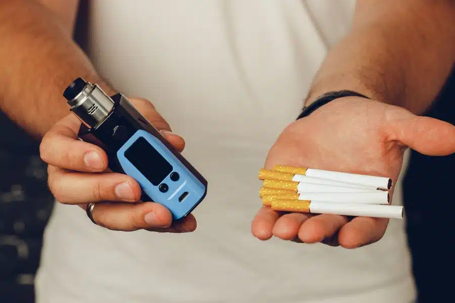 Uma mão masculina segura um vape na mão direita e seis cigarros na mão esquerda.