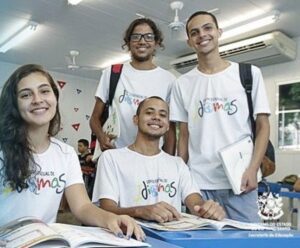 Dois jovens homens em pé, uma mulher e outro jovem hhomem sentados, com livros na mesa sorrindo