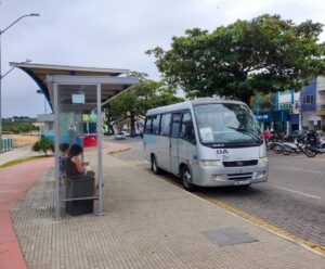 Novo sistema de transporte será tema de audiência pública em Anchieta Na imagem, uma municipe esta sentada em um ponto e espera um ônibus. A sua frente, passa um ônibus público.