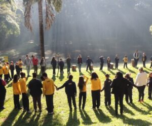 Muitas pessoas estão todas de mãos dadas, em circulo, estão em um campo abertas com coqueiros.