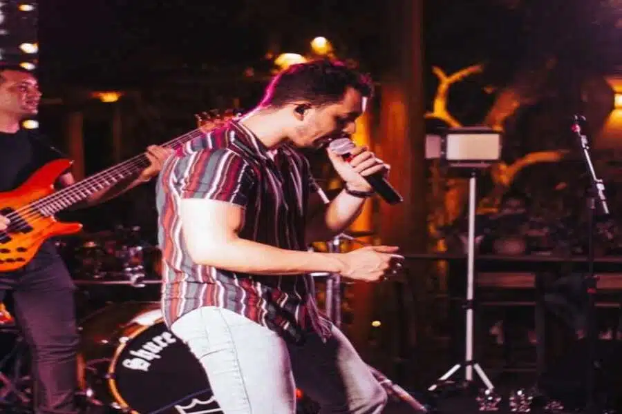 O cantor esta usando uma vamisa social, esta no palco cantando. Ao fundo os instrumentistas da banda tocam.
