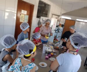 Projeto em Anchieta desenvolve novas experiências com crianças Várias crianças estão ao redor de uma mesa, com toca de cabelo e avental