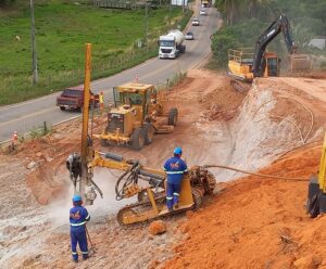 Rodovia do Frade: obras de duplicação provocam interdições Trabalhadores operam máquinas pesadas em obra de duplicação da Rodovia do Frade (ES-488), em Cachoeiro de Itapemirim, Espírito Santo. A imagem mostra operários com uniformes e equipamentos de proteção, utilizando perfuratriz em encosta de terra avermelhada. Ao fundo, escavadeira, patrol e caminhões circulam pela via parcialmente interditada com cones de sinalização.