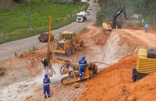Trabalhadores operam máquinas pesadas em obra de duplicação da Rodovia do Frade (ES-488), em Cachoeiro de Itapemirim, Espírito Santo. A imagem mostra operários com uniformes e equipamentos de proteção, utilizando perfuratriz em encosta de terra avermelhada. Ao fundo, escavadeira, patrol e caminhões circulam pela via parcialmente interditada com cones de sinalização.
