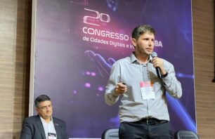 Palestrante fala ao microfone durante o 2º Congresso de Cidades Digitais e Inteligentes da Bahia. Ele está em pé, segurando um controle e usando crachá. Ao fundo, um homem sentado observa a apresentação. O painel exibe o nome do evento com elementos visuais tecnológicos.