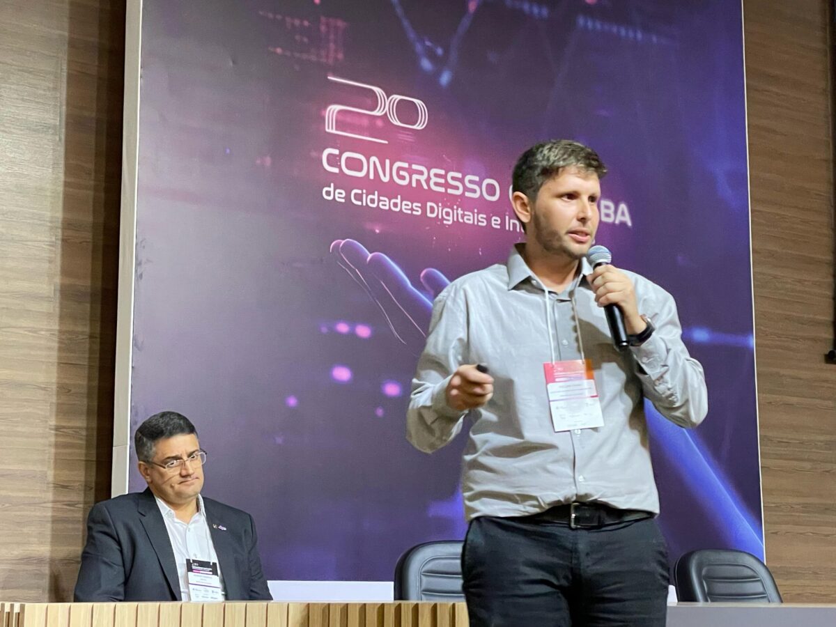 Palestrante fala ao microfone durante o 2º Congresso de Cidades Digitais e Inteligentes da Bahia. Ele está em pé, segurando um controle e usando crachá. Ao fundo, um homem sentado observa a apresentação. O painel exibe o nome do evento com elementos visuais tecnológicos.