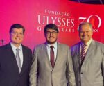 Três homens usando ternos posam para uma foto em um evento da Fundação Ulysses Guimarães, que comemora 30 anos. Ao fundo, um painel vermelho com o logotipo da fundação e a marca dos 30 anos. O ambiente está iluminado com luzes roxas, e há um palco com instrumentos musicais visíveis.