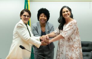 Jack Rocha é eleita coordenadora-geral da Bancada Feminina da Câmara A imagem mostra três mulheres unidas em um gesto simbólico de cooperação e força, com as mãos sobrepostas, em frente a uma bandeira do Brasil. Elas estão sorrindo, em um ambiente institucional, possivelmente uma sala de reuniões no Congresso Nacional. As vestimentas formais e o microfone na mesa indicam que o momento é parte de uma solenidade ou encontro político.