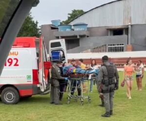 VÍDEO | Adolescente tem 25% do corpo queimado em São Roque do Canaã Equipe do SAMU e policiais militares do NOTAER realizam o transporte de um adolescente em maca até a ambulância, durante resgate aeromédico em São Roque do Canaã. Pessoas observam a ação ao fundo, em área gramada próxima a um galpão.