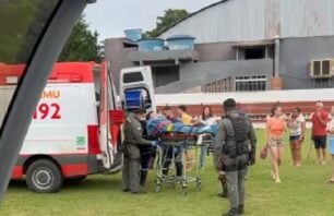 Equipe do SAMU e policiais militares do NOTAER realizam o transporte de um adolescente em maca até a ambulância, durante resgate aeromédico em São Roque do Canaã. Pessoas observam a ação ao fundo, em área gramada próxima a um galpão.