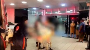 Equipes do SAMU transportam um paciente em uma maca na entrada de um hospital durante a noite, enquanto outras pessoas observam próximas à porta de emergência. A cena é iluminada pelas luzes da ambulância.