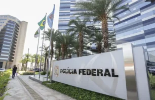 Fachada da sede da Polícia Federal em Brasília, com letreiro em destaque, bandeiras hasteadas e palmeiras alinhadas na entrada. O edifício moderno tem fachada espelhada e linhas horizontais, refletindo a paisagem ao redor. Uma mulher caminha pela calçada ao lado do prédio.