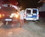 Homem de bermuda listrada e camisa xadrez, de costas, é escoltado por agentes em uma rua de paralelepípedos à noite. Ele está entre uma viatura da Polícia Civil do Espírito Santo, com luzes acesas, e uma viatura da Polícia Militar identificada como Patrulha Rural. A cena ocorre em área urbana, com casas ao fundo e iluminação artificial.