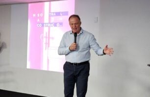 Governo abre curso de Inteligência Artificial; veja como participar Homem de camisa azul clara e calça escura fala ao microfone em um auditório, em pé diante de uma tela com projeção desfocada em tons de rosa e branco, onde se lê parcialmente "CONSTRUÇ...". Ele gesticula com a mão esquerda enquanto fala.