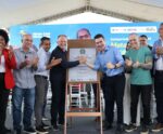 Autoridades e representantes celebram a inauguração do Residencial Mata do Cacau, do programa Minha Casa, Minha Vida, em Linhares (ES). Ao centro, duas autoridades apertam as mãos diante de uma placa comemorativa, cercadas por outras pessoas aplaudindo, sob uma tenda com painel ao fundo exibindo informações do evento.