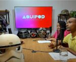 A imagem mostra uma gravação do podcast "AQUIPOD", identificado pela logomarca exibida no monitor ao fundo, com o subtítulo “O podcast do aquinoticias.com”. A cena ocorre em um estúdio decorado com capacetes e máscaras, incluindo itens inspirados em personagens como Darth Vader e Stormtrooper, da franquia Star Wars. Na mesa, há três participantes: à esquerda, uma pessoa com máscara de Darth Vader; ao centro-direita, uma mulher com blusa rosa; e à direita, um homem de camisa amarela. Todos estão posicionados diante de microfones, com papéis e copos de água à frente, indicando um ambiente de entrevista ou conversa gravada. No fundo da sala, há prateleiras com plantas e objetos decorativos, reforçando o estilo descontraído do cenário. A variedade de capacetes exibidos inclui referências à cultura pop e possivelmente a personagens como Motoqueiro Fantasma, o que pode ser tema do episódio.