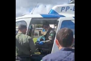 Equipe do NOTAER realiza resgate aeromédico em helicóptero. Profissionais de farda verde e azul cercam a aeronave enquanto um militar, com luvas e máscara, segura paciente no interior do veículo. A cena ocorre em área aberta durante o dia, com céu parcialmente nublado.