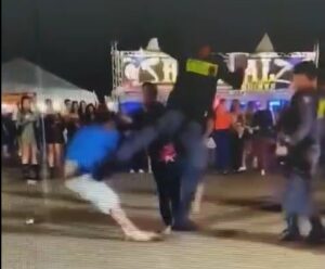 “Voadora”: policial recorre a golpe para separar briga em festa no ES A imagem mostra o momento exato em que um PM aplica um chute do tipo “voadora” em um homem de camiseta azul e bermuda clara, que parece estar caindo ao solo. O incidente ocorre em um local público, provavelmente um evento, com outras pessoas ao fundo observando a cena. Também há presença de policiais militares fardados no local, que assistem à ação.
