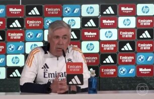 Carlo Ancelotti, treinador do Real Madrid, participa de uma coletiva de imprensa. Ele está sentado à frente de microfones, vestindo o uniforme do clube com o logotipo da Emirates. Ao fundo, há um painel com logotipos de patrocinadores como Emirates, Mahou, BMW, HP e Adidas.