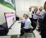 Governador e equipe técnica acompanham demonstração de teleconsulta em unidade de saúde. Um homem sentado interage com médico por videochamada exibida em tela grande, enquanto profissionais da saúde e autoridades observam a explicação. A sala é clara, com paredes brancas e decoração infantil.