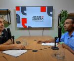A imagem mostra dois homens participando de uma entrevista ou podcast no programa "Jogo Aberto", conforme indica a tela ao fundo com a marca “Jogo Aberto – aquiéoficial.com”. Ambos estão sentados em uma mesa de madeira equipada com microfones, copos de água e documentos. O ambiente é decorado com estantes, livros e plantas.