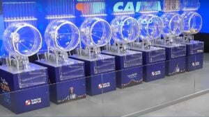 A imagem mostra os globos utilizados nos sorteios das Loterias Caixa, prontos para o sorteio da Quina de São João 2025. No centro da cena, os equipamentos transparentes e iluminados compõem o cenário tradicional dos concursos especiais, com destaque para o logotipo das Loterias Caixa e elementos gráficos típicos das festas juninas. O palco já está preparado para a extração que promete pagar o maior prêmio da história da Quina de São João, estimado em R$ 250 milhões.