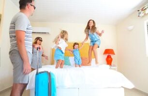 Família com crianças se diverte em quarto de hotel durante viagem, com malas no chão e clima de alegria e descontração