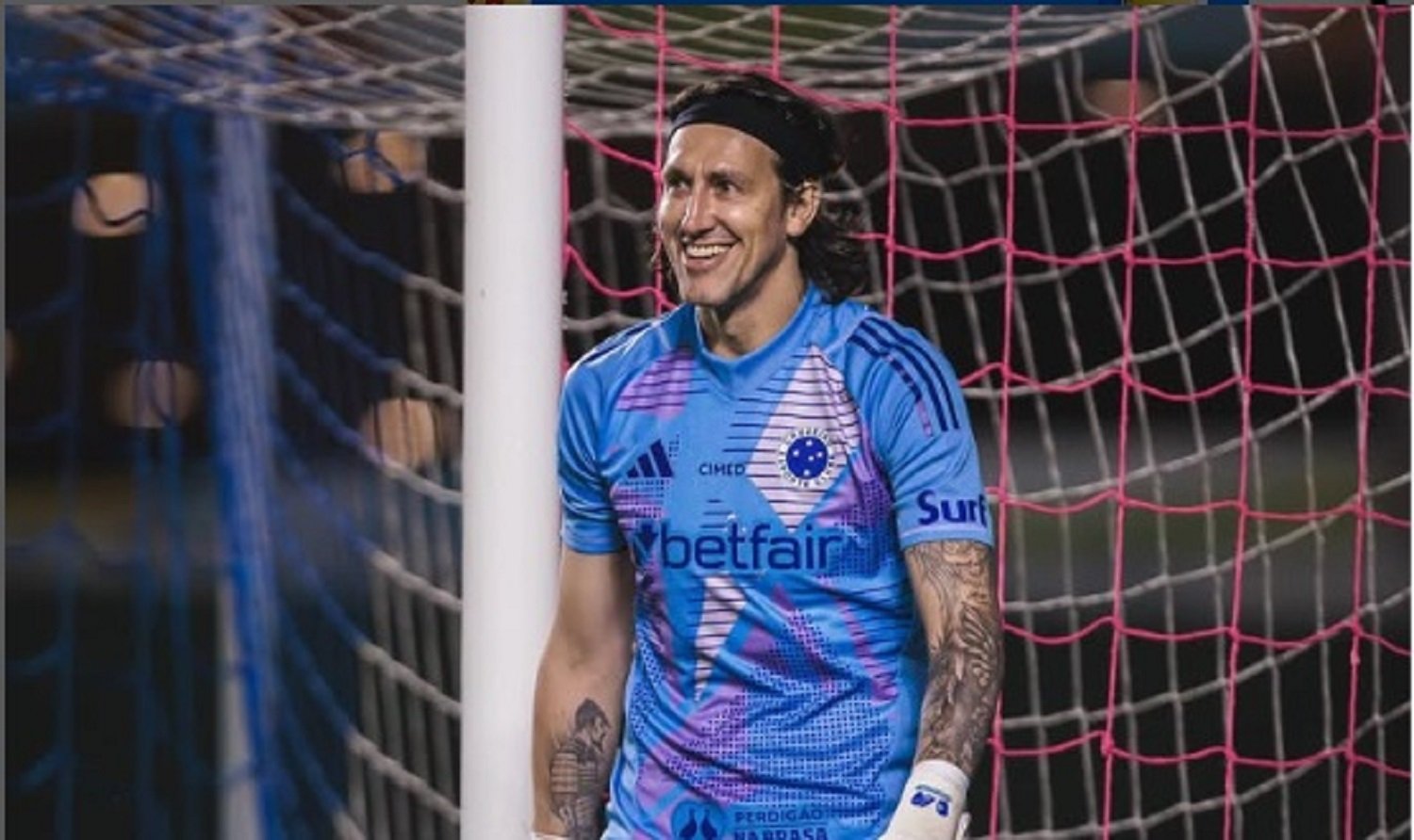 A imagem mostra um goleiro sorrindo, posicionado à frente de uma trave com redes nas cores rosa e azul. Ele veste um uniforme azul com detalhes em roxo e rosa, que inclui a logomarca do patrocinador "Betfair" e o escudo do Cruzeiro Esporte Clube. O atleta também usa luvas de goleiro, uma faixa preta na cabeça e possui tatuagens no braço esquerdo.