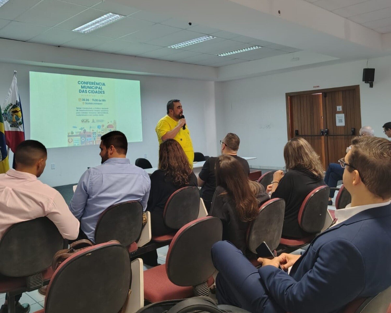 Homem de camisa amarela fala ao microfone durante a Conferência Municipal das Cidades, com plateia atenta em auditório e apresentação projetada ao fundo.