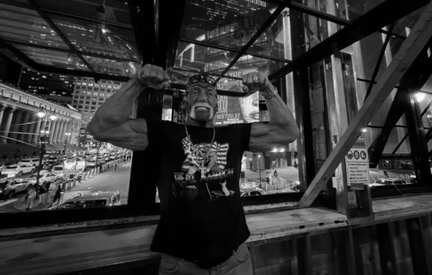 Hulk Hogan, com camiseta estampada e bandana, posa mostrando os músculos em frente a uma janela com vista para uma avenida movimentada à noite. A imagem em preto e branco destaca sua expressão sorridente e a iluminação urbana ao fundo.
