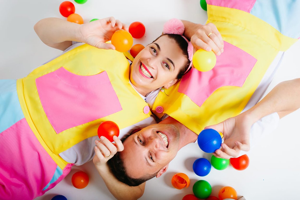 Dois adultos sorridentes, vestidos com aventais coloridos em amarelo, rosa e azul, deitados no chão cercados por bolas de plástico multicoloridas, transmitindo clima alegre e divertido.