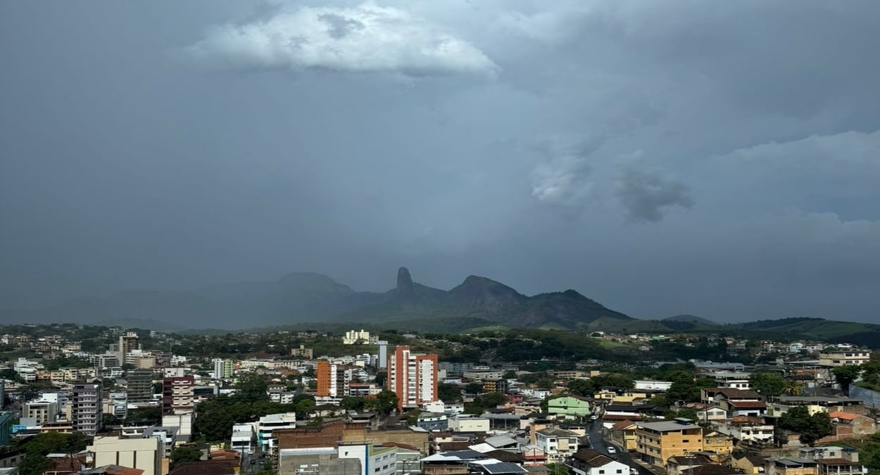 Panorama de Cachoeiro de Itapemirim durante chuva forte em Cachoeiro, com nuvens escuras avançando sobre a cidade e a Pedra do Itapemirim ao fundo.