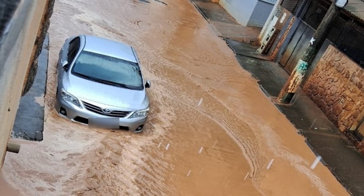 Rua alagada no bairro São Geraldo após chuva forte em Cachoeiro, com carro ilhado no meio da água barrenta.