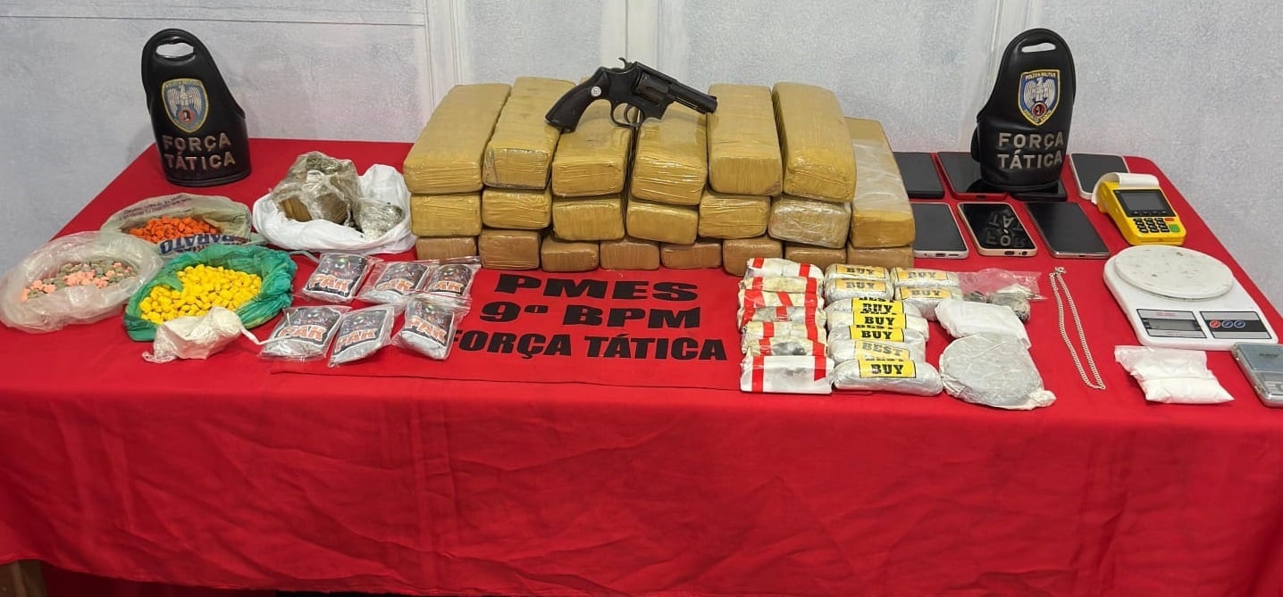 Mesa coberta por um pano vermelho exibe grande quantidade de drogas apreendidas, incluindo dezenas de tabletes de maconha, comprimidos de ecstasy, porções de cocaína, embalagens diversas, duas armas de fogo, sete celulares, balanças de precisão e máquinas de cartão. Ao fundo, há dois escudos da Força Tática da PMES identificando a operação do 9º BPM.