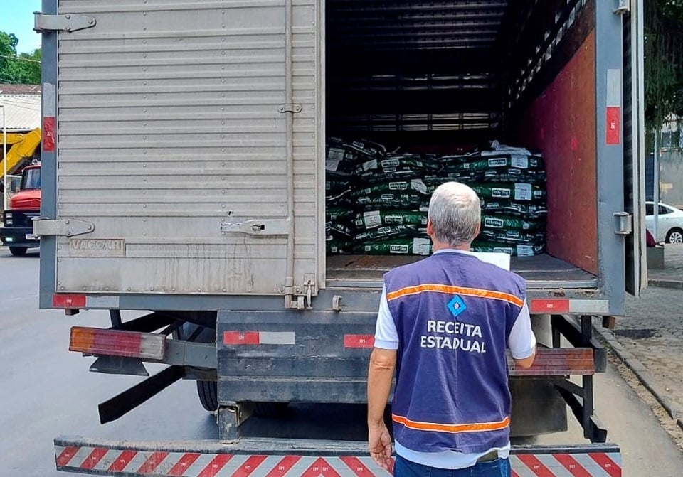 Auditor da Receita Estadual observa o interior de um caminhão de carga aberto durante uma fiscalização. O veículo transporta diversos sacos empilhados, enquanto o agente, usando colete identificador, verifica a mercadoria em uma rua movimentada.