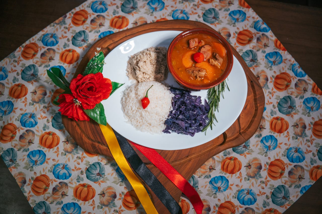 Prato de feijoada alemã servido com arroz, repolho roxo refogado e farofa, acompanhado de decoração com flor vermelha e fitas coloridas sobre uma tábua de madeira.