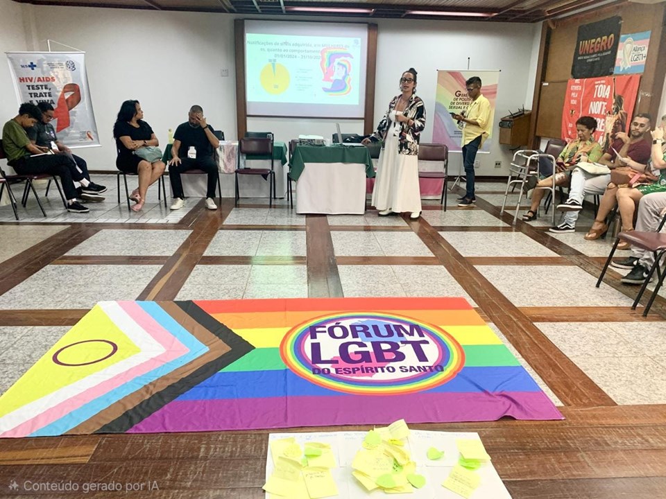 Pessoas participam de um encontro sobre saúde e cidadania LGBTI+ em sala ampla, com bandeira do Fórum LGBT do Espírito Santo estendida no chão, apresentação em tela ao fundo e participantes sentados em roda.