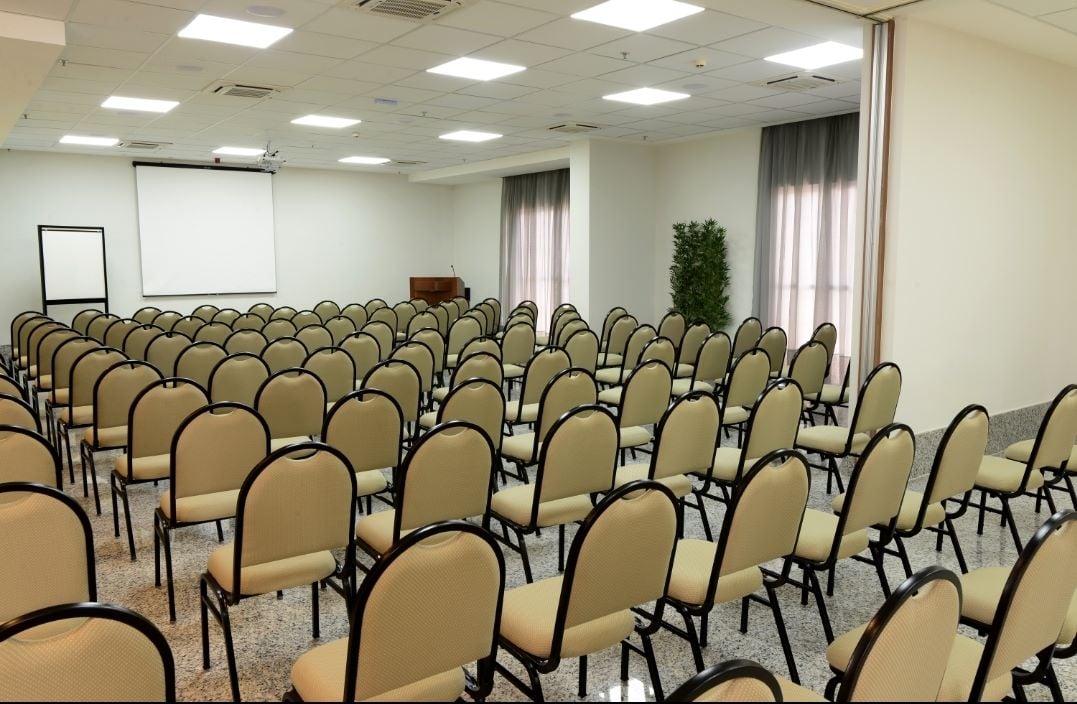 Sala de eventos ampla e iluminada, com várias fileiras de cadeiras organizadas em formato auditório, tela de projeção ao fundo e cortinas claras nas janelas, pronta para receber palestras ou reuniões corporativas.