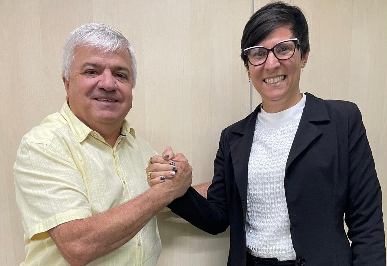 Mulher sorridente de óculos e blazer preto cumprimenta com as mãos um homem de camisa amarela, ambos posando lado a lado em frente a uma parede clara.